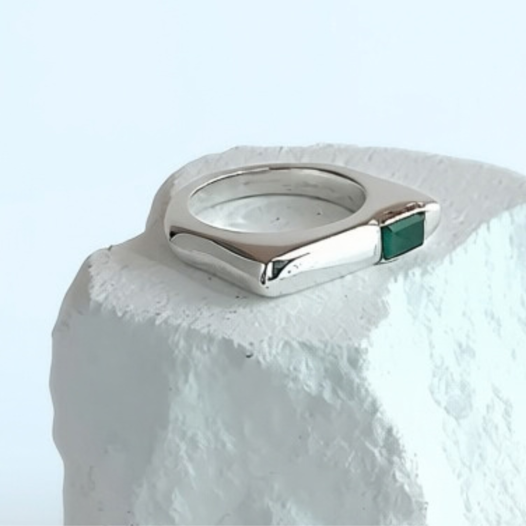 A silver-toned geometric ring with a small green rectangular stone, displayed on a textured white block against a light blue background.